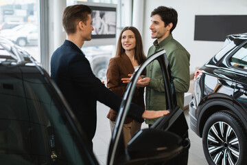Joyful young couple consulting with auto salesman, picking new car at automobile dealership