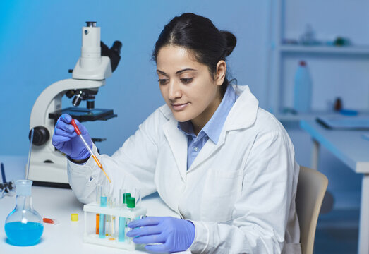 Content Indian female clinician in lab coat and rubber gloves sitting at desk and using colorful reagents while carrying out chemical experiment in laboratory