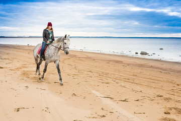 dressing jeans, jacket and autumn hat female jokey rides astraddle a dappled horse along seaside
