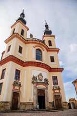 Fototapeta premium Litomysl, Czech Republic, 17 April 2022: Church of the Finding of the Holy Cross and Piarist dormitory near castle, baroque building with tower at sunny summer day, stone statues in the garden
