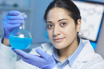Fototapeta premium Portrait of content confident young mixed race female chemist in blue protective gloves holding laboratory flask of cyan liquid