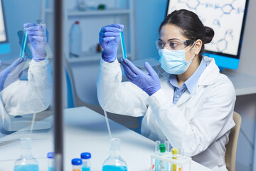 Serious young Indian female chemist in lab coat and surgical mask sitting at desk and making experiment with liquid substances