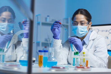 Concentrated young Indian female laboratory worker in mask and safety goggles sitting at desk and adding blue liquid into test tube