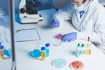 Busy lab worker in white coat and mask sitting at table with medical substances and analyzing bacteria sample in petri dish