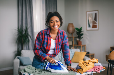 Smiling pretty young african american woman housewife in casual ironing clothes with iron in living...