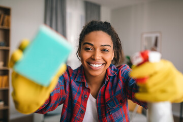 Smiling young african american lady in rubber gloves with spray and sponge wipes camera at modern kitchen - obrazy, fototapety, plakaty
