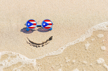 Fototapeta na wymiar A painted smile on the beach and sunglasses with the flag of the Puerto Rico. The concept of a positive holiday in the resort of the Puerto Rico.