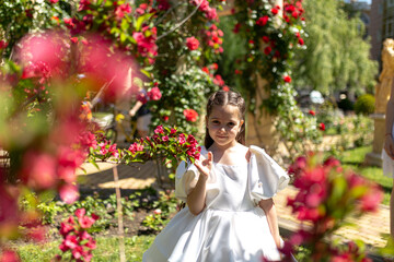 Summer portrait of pretty little girl wearing white dress, posing in red rose garden smelling flowers. Happy smiling girl