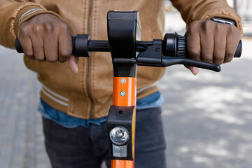 Close up of hands of an African-American man are located on the steering of the electric scooter. Dark-skinned man in a fashionable jacket professionally drives an urban electric scooter for travel