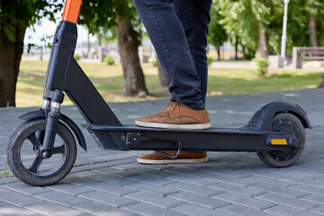 Obraz premium Close up of men's legs ready to start in a scooter race. Against the background of trees in a city park, a photo of a modern electric scooter and a man riding on paving slabs