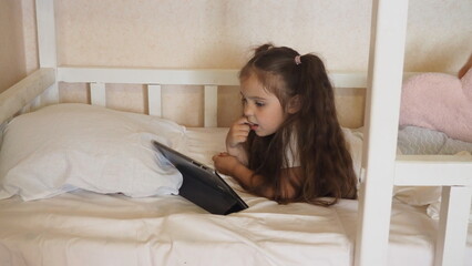 Obraz na płótnie Canvas Little girl with tablet and headphones on the bed at home. Child girl listening to music or watching cartoons, chatting with friends or family before bed