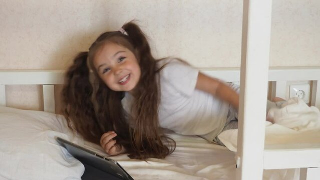 Little girl with tablet and headphones on the bed at home. Child girl listening to music or watching cartoons, chatting with friends or family before bed