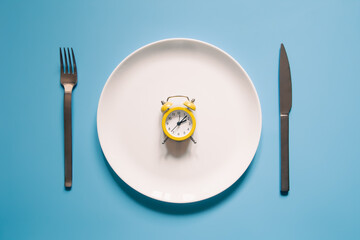 Alarm clock on a white plate with a knife and fork on blue background.