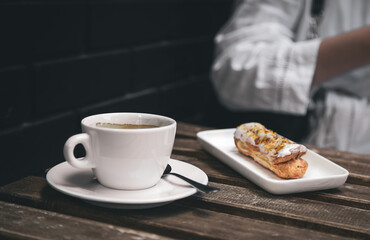 Cup of coffee and eclair on a wooden table close-up.