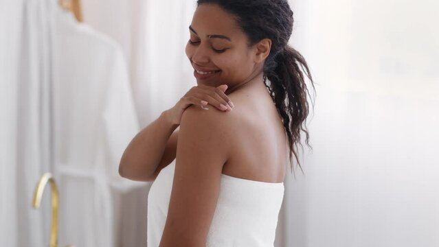 Self pampering concept. Beautiful black lady wrapped in towel applying body cream on shoulder after bath at home