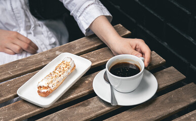 Cup of coffee and eclair on a wooden table close-up.