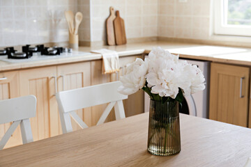Beautiful peony flowers in glass vase on the kitchen table. Close up shot of a lush bouquet in stylish modern kitchen with small beige tile and wooden cupboard. Close up, copy space, background.