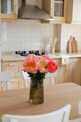 Beautiful peony flowers in glass vase on the kitchen table. Close up shot of a lush bouquet in stylish modern kitchen with small beige tile and wooden cupboard. Close up, copy space, background.