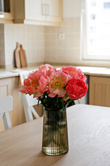Beautiful peony flowers in glass vase on the kitchen table. Close up shot of a lush bouquet in stylish modern kitchen with small beige tile and wooden cupboard. Close up, copy space, background.