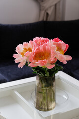 Close up shot of glass coffee table with bouquet of beautiful pink peony flowers in a vase and blue textile couch under the window on the background. Copy space, close up, natural light.
