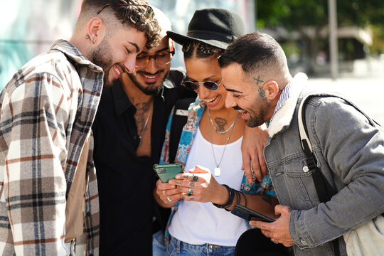 Happy group of friends watching social media on screen mobile phone, laughing. Young people using smartphone, having fun together in the city.
