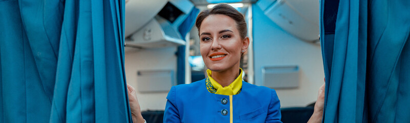 Joyful flight attendant in air hostess uniform looking at camera and smiling while holding drapes...