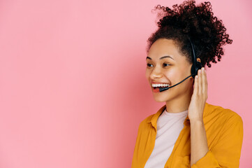 Close-up of positive african american curly haired young woman with headset, help desk worker, call center operator, consultant, talking with client, stand on isolated pink background, smiles friendly - obrazy, fototapety, plakaty