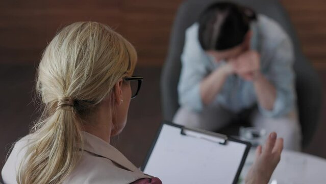 Over shoulder view of female psychologist sitting in armchair, talking with upset woman patient. Psychologist taking notes on clipboard. Psychology, mental therapy, mental health, therapy session.