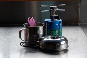 A small, compact kettle sits in front of a tin mug with a plastic spork sitting in it. It is next to the small gas burner.