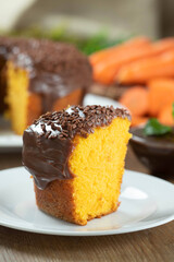 Close up piece of Brazilian carrot cake with chocolate frosting on wooden table with carrots in the background