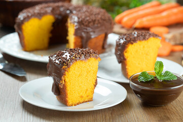 Close up piece of Brazilian carrot cake with chocolate frosting on wooden table with carrots in the background