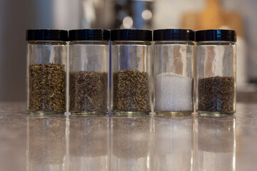 Side view of five spice bottles, salt, cilantro, italian seasoning, crushed mint, dill with blurred background