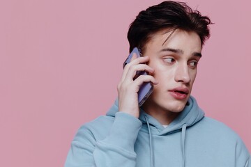 a handsome, thoughtful man is standing on a pink background in a light blue hoodie and holding a smartphone in his hands talking on it. Horizontal Studio Photography