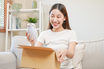 Happy excited, asian young woman, girl customer sitting on sofa at home, opening and unpacking cardboard box carton parcel after buying ordering present, shopping online, delivery service concept. - obrazy, fototapety, plakaty