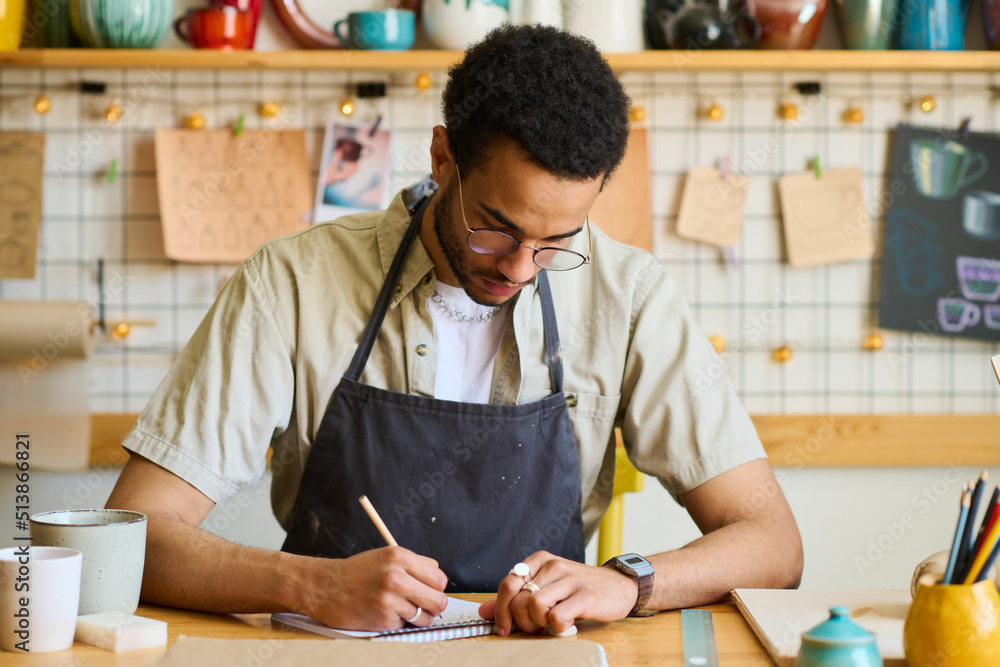 Poster young man in casual shirt and black apron drawing sketch of clay item in notepad while working over 