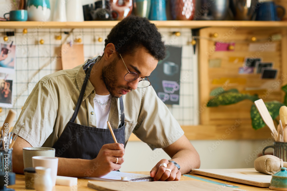 Poster creative black man in workwear and eyeglasses bending over table while looking at page of notepad wi