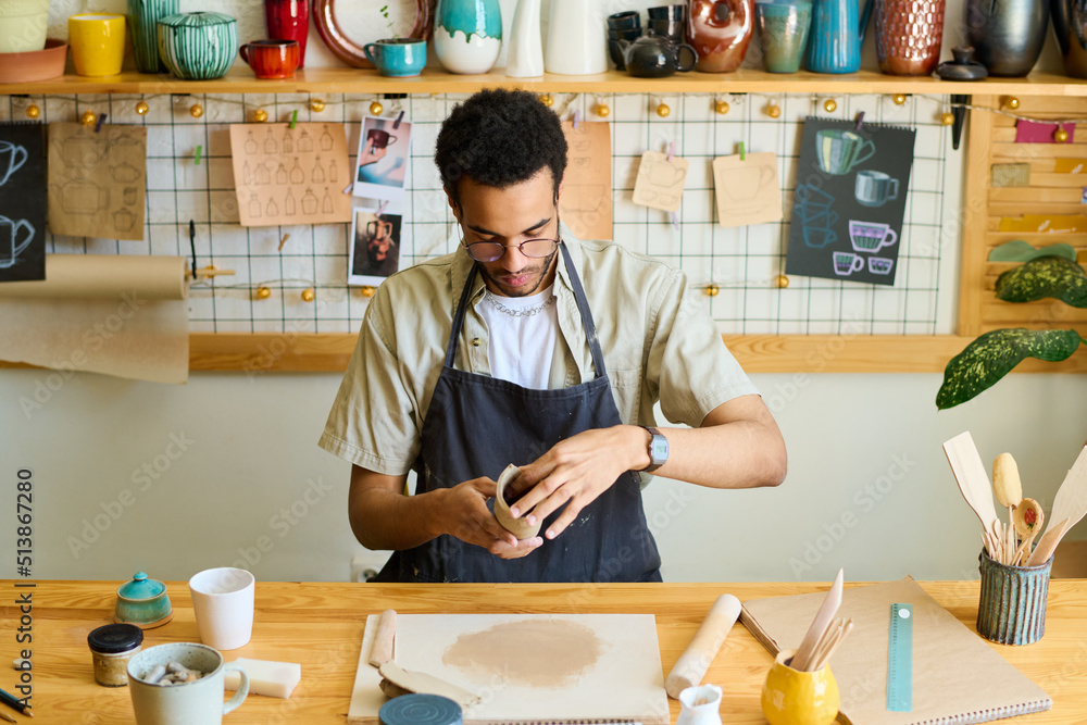 Aufkleber Young creative black man in workwear creating new clay item for sale while sitting by table against shelf with earthenware in workshop