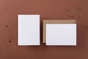 Blank greeting cards and envelopes with dry flowers on brown background. Blank paper sheet cards with mockup copy space. Minimal workplace composition. Flat lay, top view