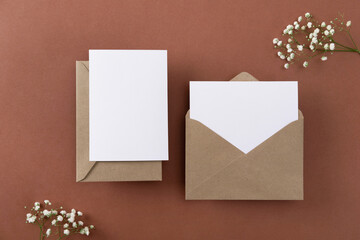 Blank greeting cards and envelopes with dry flowers on brown background. Blank paper sheet cards with mockup copy space. Minimal workplace composition. Flat lay, top view