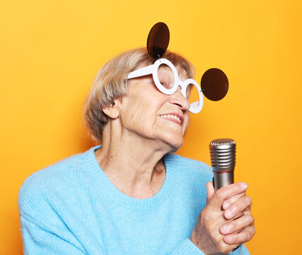 Happy Old Woman Singing With Microphone Over Yellow Color Background, Having Fun