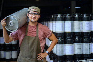 Male brewer carrying metal beer kegs at brewery Small Business and the Beer Industry A smiling handsome young man in an apron holds a steel beer keg on his shoulder in a warehouse. - Powered by Adobe
