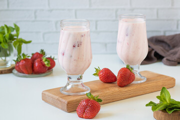 Milkshake in glasses on a wooden stand. A dish with strawberries and mint in the background. 
