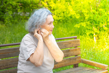 A gray-haired elderly woman adjusts her earring while sitting on a park bench