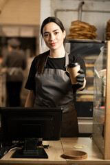 happy saleswoman with paper coffee cup giving americano coffee in vegan cafe. small business, takeaway food, people and service