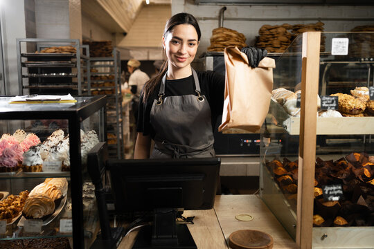 baker woman standing with fresh bread and pastry at bakery. Young woman in her bake shop looking at camera. baker with breads in background. Girl owner bakery shop