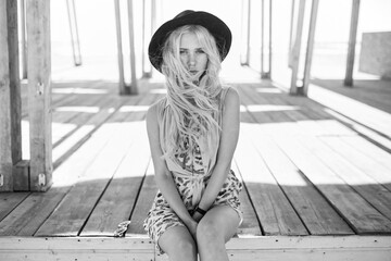 Charming blonde with curly hair, in a dress and hat, isolated at the wooden building background. Black and white photo.
