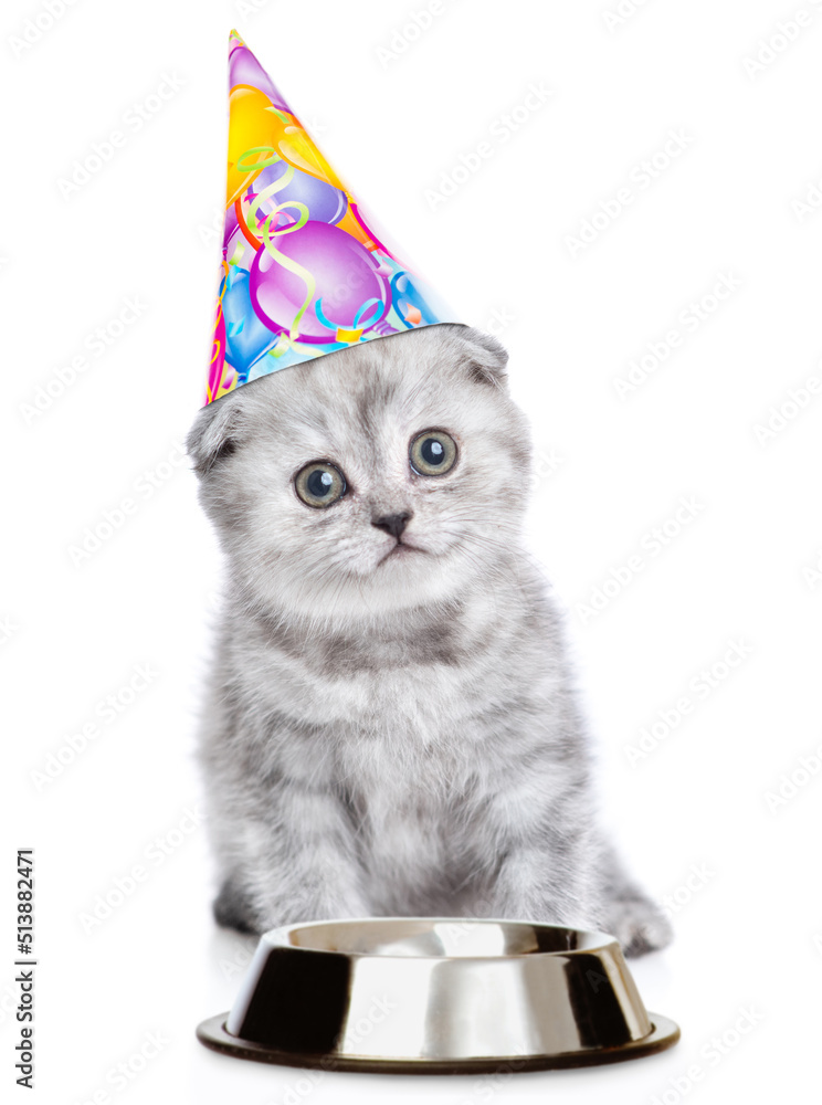 Poster cute kitten wearing party cap sits with empty bowl and looks at camera. isolated on white background