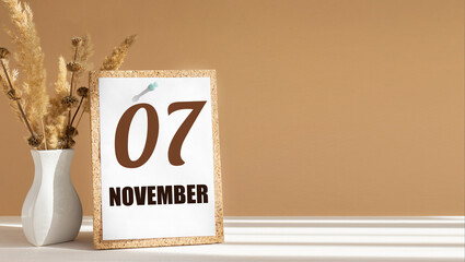 november 7. 7th day of month, calendar date.White vase with dead wood next to cork board with numbers. White-beige background with striped shadow. Concept of day of year, time planner, autumn month