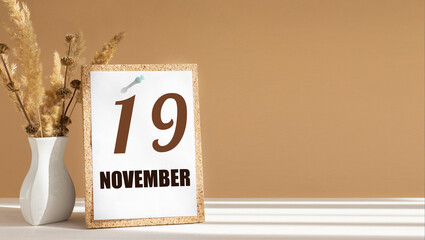november 19. 19th day of month, calendar date.White vase with dead wood next to cork board with numbers. White-beige background with striped shadow. Concept of day of year, time planner, autumn month