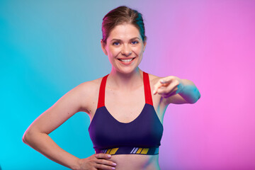 Coach Girl points a finger straight in front of her. Smiling sporty woman in fitness bra sportswear. Female fitness portrait isolated on neon multicolor background.